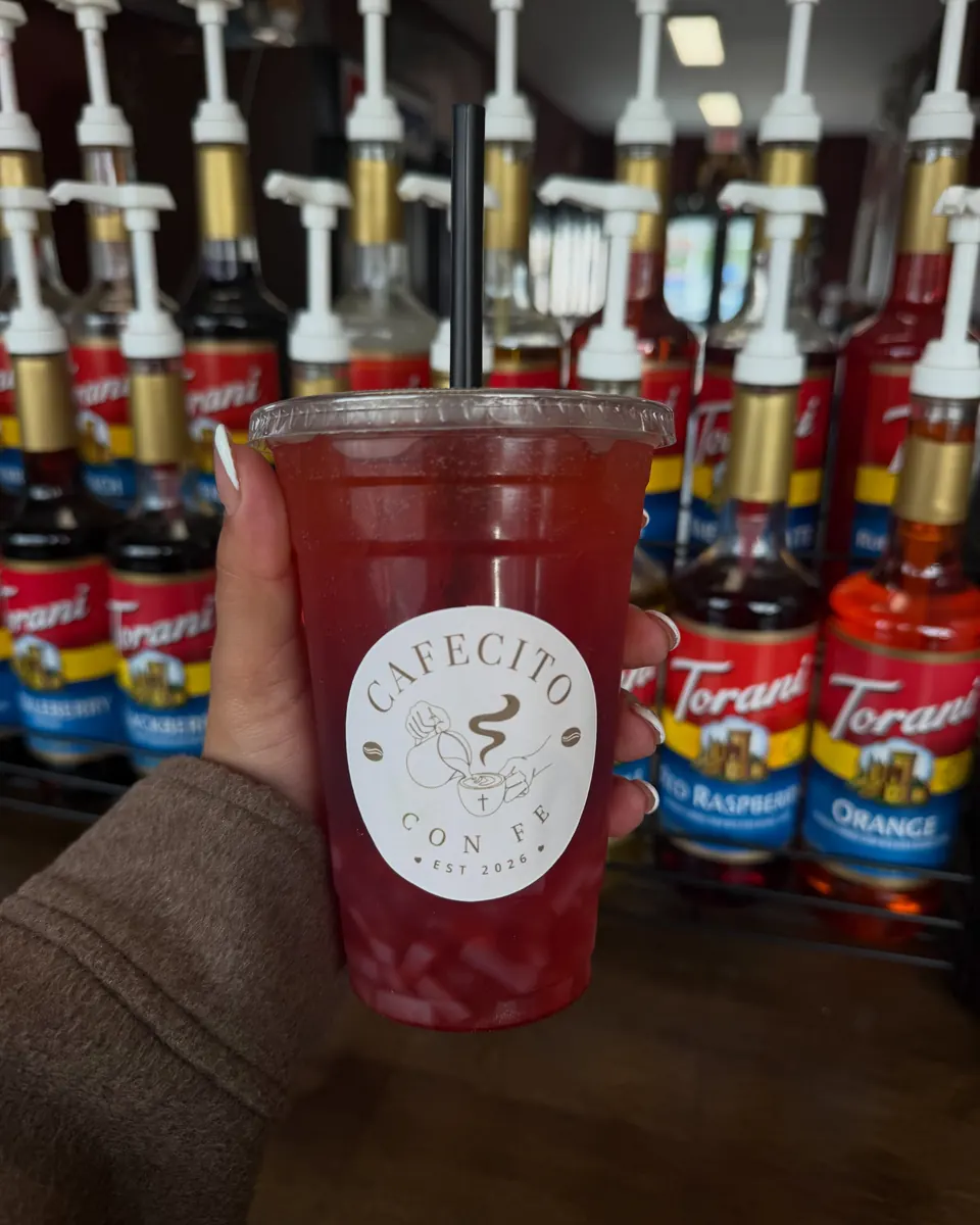 Red Cafecito Con Fe drink held in front of the Torani syrup wall at the counter